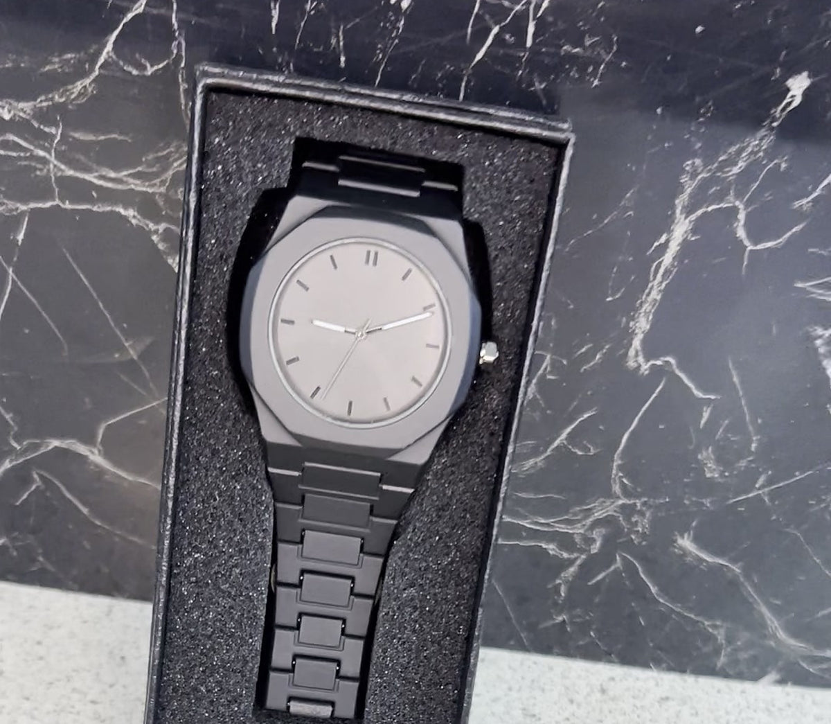 Reloj negro mate liso con esfera gris grafito e índices de barra plateados, dentro de su caja negra con espuma, sobre mármol (foto real)